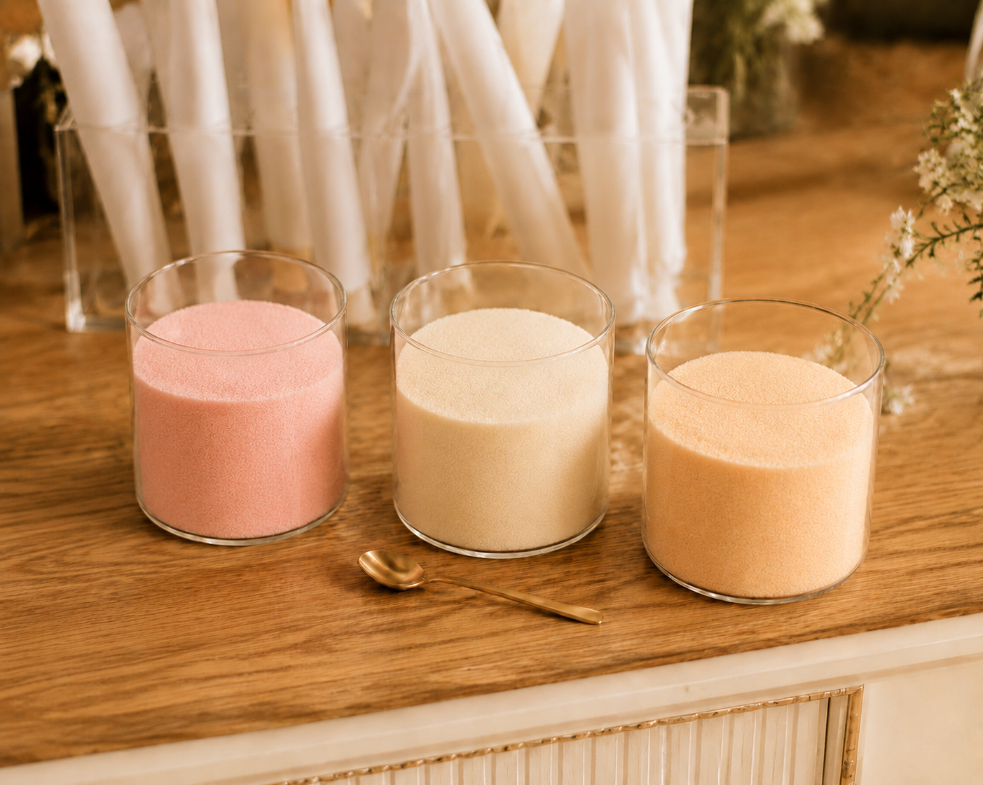 Three glass jars of blush, cream, and peach pure cane sugar on a wooden cart surface with a small gold spoon