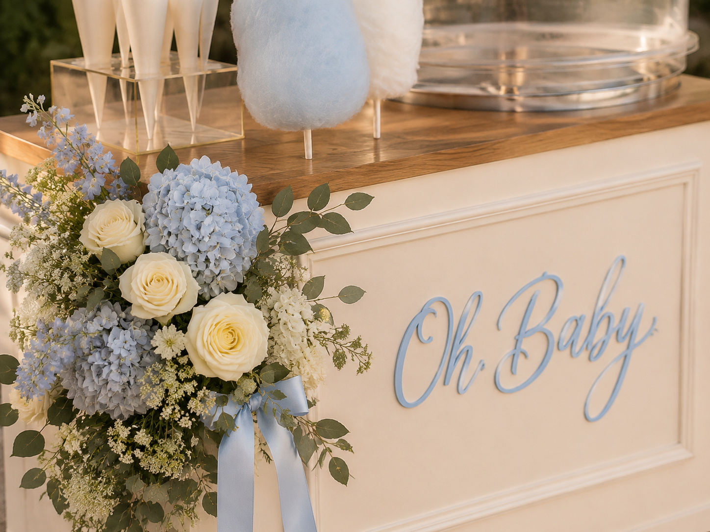 Ivory cotton candy cart styled with blue hydrangeas, cream roses, and custom Oh Baby signage